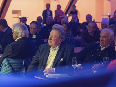 Gäste an Tischen im Saal blicken zur Bühne, vorn ein Mann mit schachbrettgemusterter Fliege und Einstecktuch