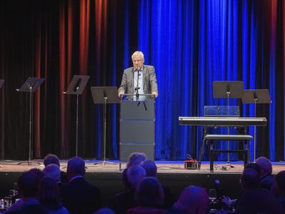 Redner am Pult zwischen Notenpulten und Digitalpiano vor blauem Vorhang, im Vordergrund Publikum an Tischen