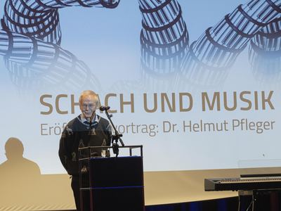 Festredner im Pullover mit Schachfiguren-Muster am Pult vor der Projektion „Schach und Musik“ mit spiralförmigen Klaviertasten