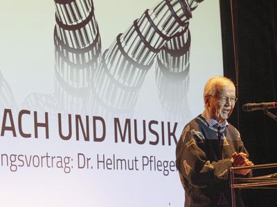 Festredner im Schachfiguren-Pullover spricht am Pult, hinter ihm die Titelprojektion „Schach und Musik – Eröffnungsvortrag“