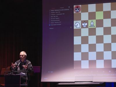 Vortragender am Pult, daneben auf der Leinwand ein großes Schachdiagramm mit Mattstellung und eine Liste historischer Partien