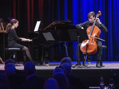 Junge Pianistin am Flügel und junger Cellist spielen gemeinsam auf der blau beleuchteten Bühne, vorn Zuschauer