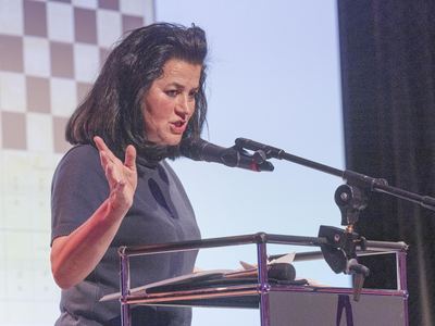 Rednerin stellt am Pult den Ausstellungskatalog vor, hinter ihr eine Projektion mit Schachbrettmuster und Notenlinien