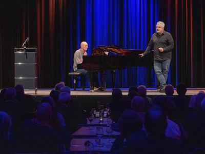 Sänger und Pianist am Flügel auf der Bühne, im Vordergrund Gäste an Tischen mit Gläsern und Teelichtern