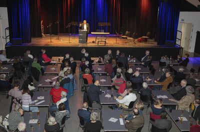 Blick von oben in den Saal: Gäste an Tischen, auf der Bühne ein Redner am Pult zwischen Notenpulten und Instrumenten