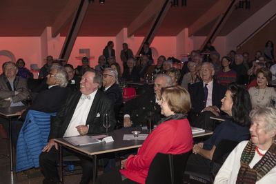 Lachendes Publikum an Tischen im Saal mit Holzdachbalken, vorn ein Gast mit schachgemusterter Fliege