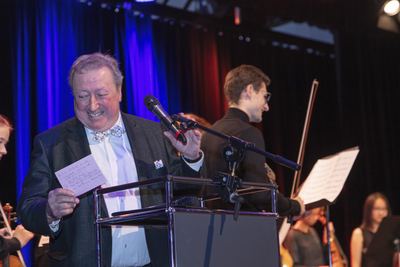 Lächelnder Moderator mit Karteikarte am Rednerpult, hinter ihm Musikerinnen und Musiker des Orchesters mit Noten