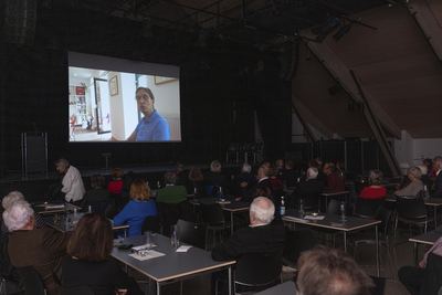 Filmvorführung im abgedunkelten Saal: Auf der Leinwand spricht ein Mann in einem Raum mit großen Schachfiguren, Gäste schauen zu