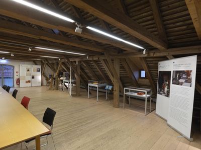Ausstellungsraum unter einem alten Holzdachstuhl mit Vitrinen, Schachspielen, Notenpult und großen Texttafeln zu einem Schachgemälde