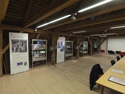 Dachgeschoss mit Ausstellungsbannern und Glasvitrinen voller Schallplatten mit Schachmotiven, rechts Stuhlreihen vor einer Leinwand