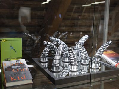 Skulptur aus gewundenen Röhren mit Klaviertastenmuster auf einem Spiegel in einer Vitrine, daneben Bücher zu Musik und Schach