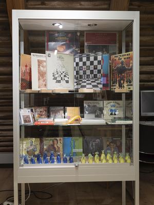 Hohe Glasvitrine mit klassischen Schallplatten mit Schachmotiven, Liederheften und einem blau-gelben Schachspiel aus Keramik