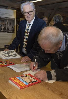 Ein Gast signiert ein Schachbuch am Holztisch, neben ihm ein Mann mit Schachkrawatte und der Ausstellungskatalog „Schach und Musik“