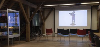 Stuhlreihen vor einer Leinwand mit der Projektion einer Porzellanfigur eines Klarinettisten, links eine Vitrine und eine Tafel mit Noten