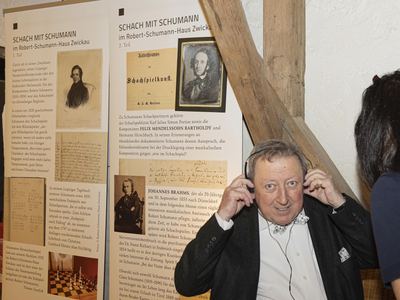 Gast mit Fliege setzt sich Kopfhörer auf, hinter ihm Ausstellungstafeln über einen Komponisten und sein Schachspiel