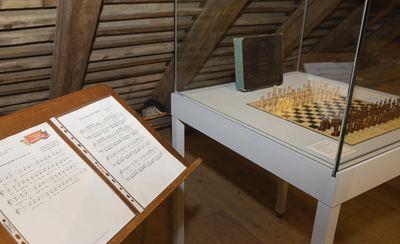 Notenpult mit Schachliedern im Vordergrund, dahinter eine Vitrine mit historischem Schachspiel und altem Buch