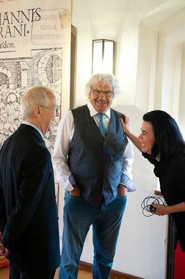 Drei Gäste der Eröffnung lachen im Gespräch vor einem großformatigen alten Holzschnitt-Titelblatt