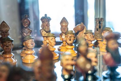 Geschnitzte Holzschachfiguren mit karikaturhaften Gesichtern: König, Dame, Läufer mit Mitra und Bauern mit Hut in einer Vitrine