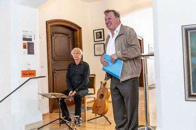 Ein Redner mit blauer Mappe begrüßt die Gäste im Rathausfoyer, hinter ihm sitzt ein Gitarrist neben Notenständer und Gitarre