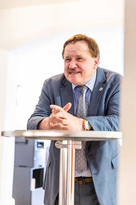Ein Redner im grauen Anzug stützt sich mit gefalteten Händen auf einen Stehtisch und spricht zum Publikum