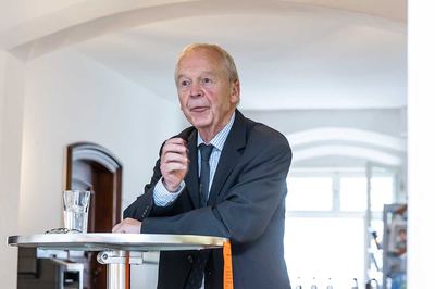 Ein älterer Vortragender im dunklen Anzug steht am Stehtisch mit Wasserglas und spricht zum Publikum