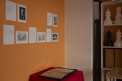 Ausstellungsecke mit Porträts, Zeichnungen und Texttafeln an oranger Wand, davor ein Ordner auf rotem Tisch und große Schachfiguren im Regal