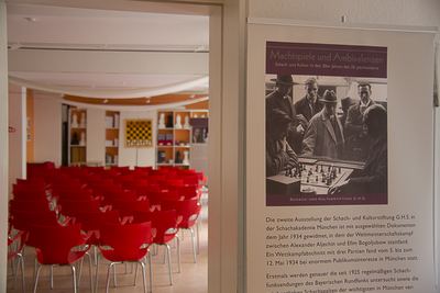 Plakat der Ausstellung „Machtspiele und Ambivalenzen“ mit historischem Foto einer umlagerten Schachpartie, links der Saal mit roten Stühlen