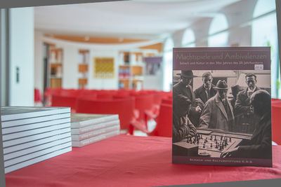 Katalog „Machtspiele und Ambivalenzen“ aufgestellt auf rot gedecktem Tisch neben Bücherstapeln, dahinter der Saal der Schachakademie