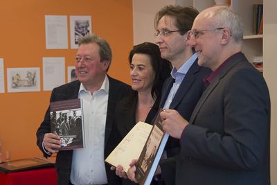 Vier Personen vor der orangen Ausstellungswand halten den Katalog und das aufgeschlagene historische Gästebuch in die Kamera