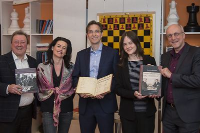 Fünf Personen vor einem großen Wandschachbrett, in den Händen Kataloge, ein Schachbuch und das aufgeschlagene alte Gästebuch