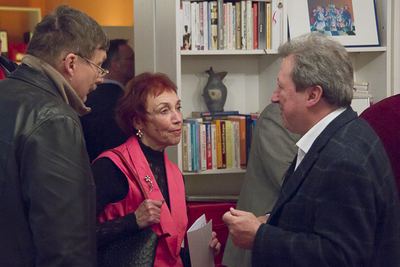 Drei Gäste der Buchpräsentation im Gespräch vor einem Bücherregal mit Vase und gerahmtem Bild
