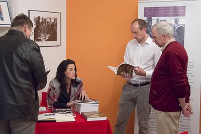 Zwei Besucher blättern im Katalog neben dem Büchertisch, dahinter das Ausstellungsbanner „Machtspiele und Ambivalenzen“