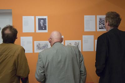 Drei Besucher von hinten betrachten Porträts, Zeichnungen und Texttafeln zum Münchner Schachleben 1934 an oranger Wand