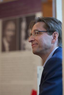 Mann mit Brille im Profil, lächelnd, im unscharfen Hintergrund ein Ausstellungsbanner mit Schwarzweißporträts
