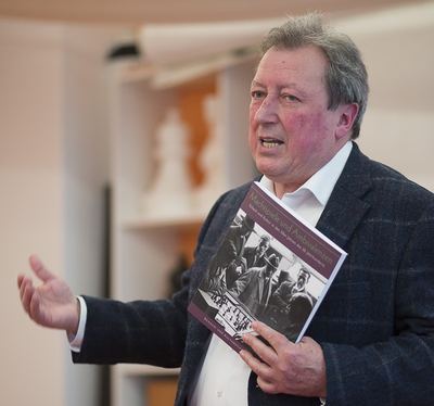 Redner präsentiert gestikulierend den Katalog „Machtspiele und Ambivalenzen“ mit historischem Schachfoto auf dem Umschlag