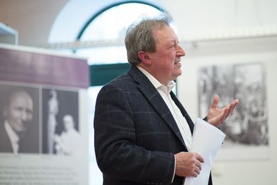 Redner mit Papieren in der Hand spricht vor einem Rundbogenfenster, links ein Ausstellungsbanner mit Schwarzweißporträts