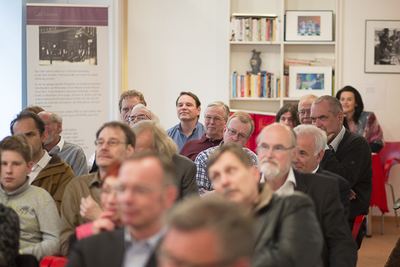 Zuhörer in Stuhlreihen verfolgen die Buchpräsentation, dahinter ein Ausstellungsbanner mit historischem Foto und Bücherregal