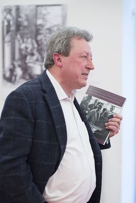 Redner im Profil hält den Katalog „Machtspiele und Ambivalenzen“, im Hintergrund ein historischer Kupferstich