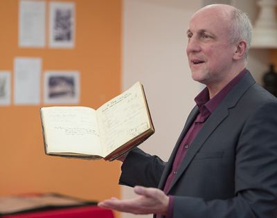 Redner zeigt dem Publikum das aufgeschlagene Gästebuch mit handschriftlichen Einträgen, hinten Ausstellungstafeln an oranger Wand