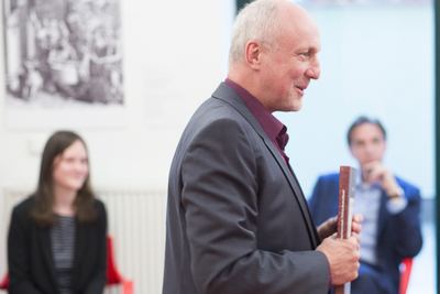 Mann mit einem Buch in der Hand im Profil, dahinter unscharf zwei Zuhörer und ein historischer Kupferstich an der Wand
