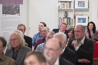 Aufmerksame Zuhörer in Stuhlreihen, links ein Ausstellungsbanner zum Münchner Café Stefanie mit historischem Foto