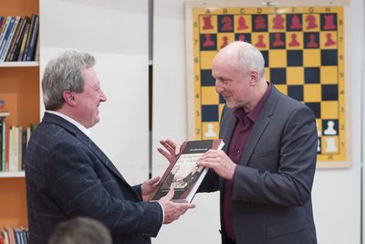 Zwei Männer überreichen einander lächelnd ein Buch, hinter ihnen das große Wandschachbrett