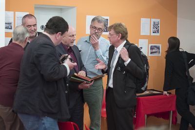 Gruppe von Gästen begutachtet das historische Gästebuch vor der orangen Ausstellungswand mit gerahmten Fotos