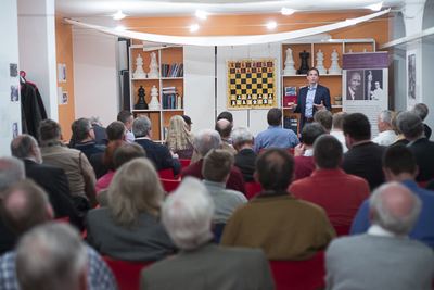 Gut gefüllter Saal der Schachakademie: Ein Redner spricht vor Wandschachbrett und großen Schachfiguren zum Publikum