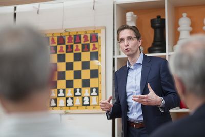 Redner mit Brille erklärt gestikulierend vor einem großen Wandschachbrett mit Demonstrationsfiguren