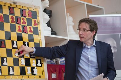 Redner zeigt auf eine Figur am Wandschachbrett, um eine Stellung zu erläutern, dahinter Regal mit großen Schachfiguren
