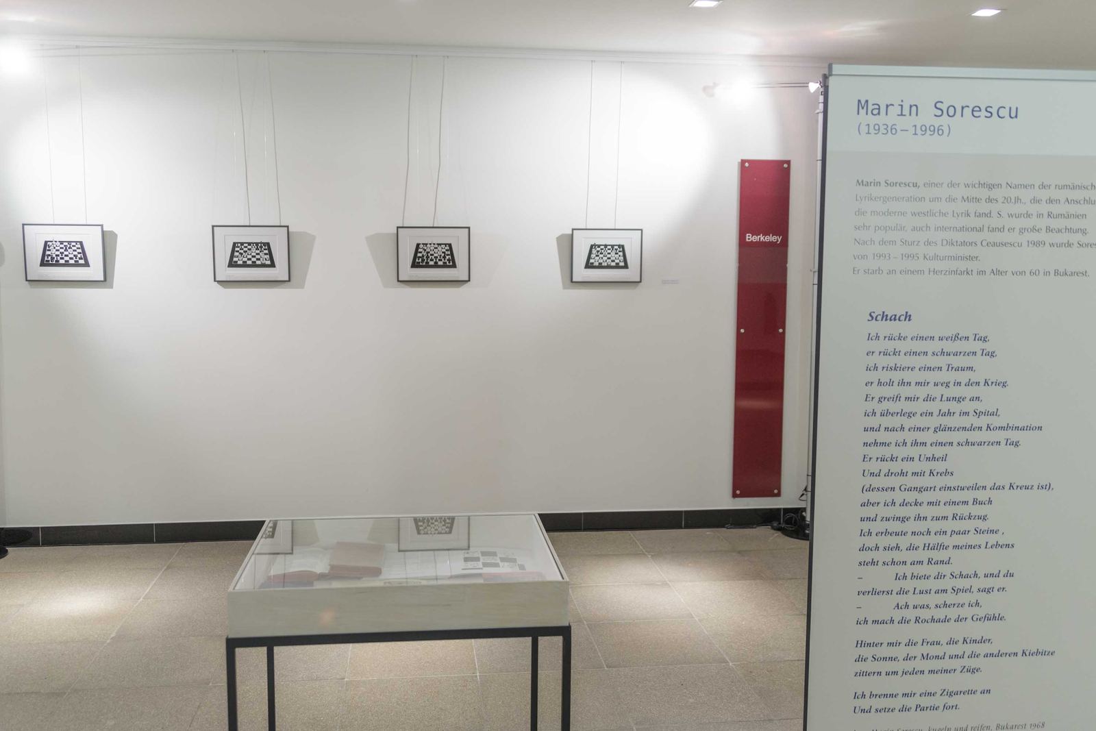 Aufgestellte Texttafel mit Kurzbiografie und dem Gedicht „Schach“ vor einer Wand mit vier gerahmten Schachbrett-Grafiken und einer Vitrine.