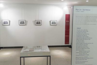 Aufgestellte Texttafel mit Kurzbiografie und dem Gedicht „Schach“ vor einer Wand mit vier gerahmten Schachbrett-Grafiken und einer Vitrine.