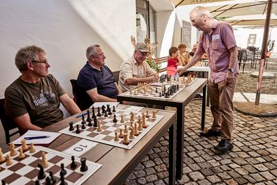Großmeister beugt sich beim Simultanspiel über ein Brett, die Gegner sitzen aufgereiht an Tischen unter Sonnenschirmen