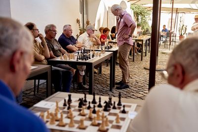 Blick über ein Schachbrett auf die Tischreihen im Hof, der Großmeister überlegt stehend mit der Hand am Kinn